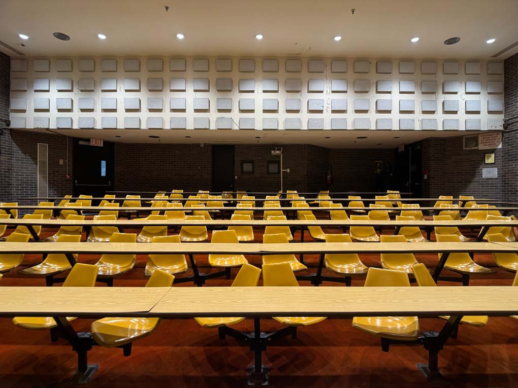 Tiered seating lecture hall. Photo of an empty tiered seating lecture hall with yellow seats and red carpet.