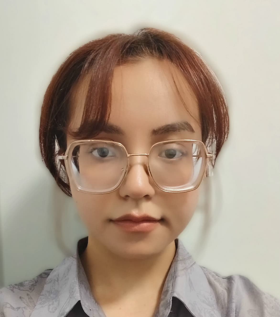 Young woman with tied back reddish hair and bangs, wearing glasses and a grey collared shirt against a beige wall