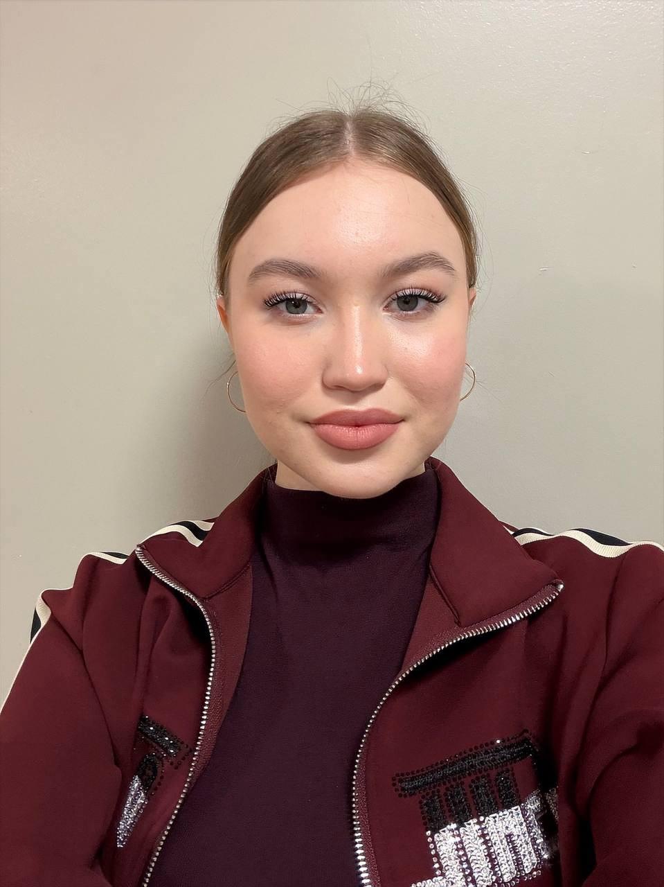 Photo of a young woman with tied back blonde hair wearing a burgundy high neck top and a track suit jacket against a beige wall.