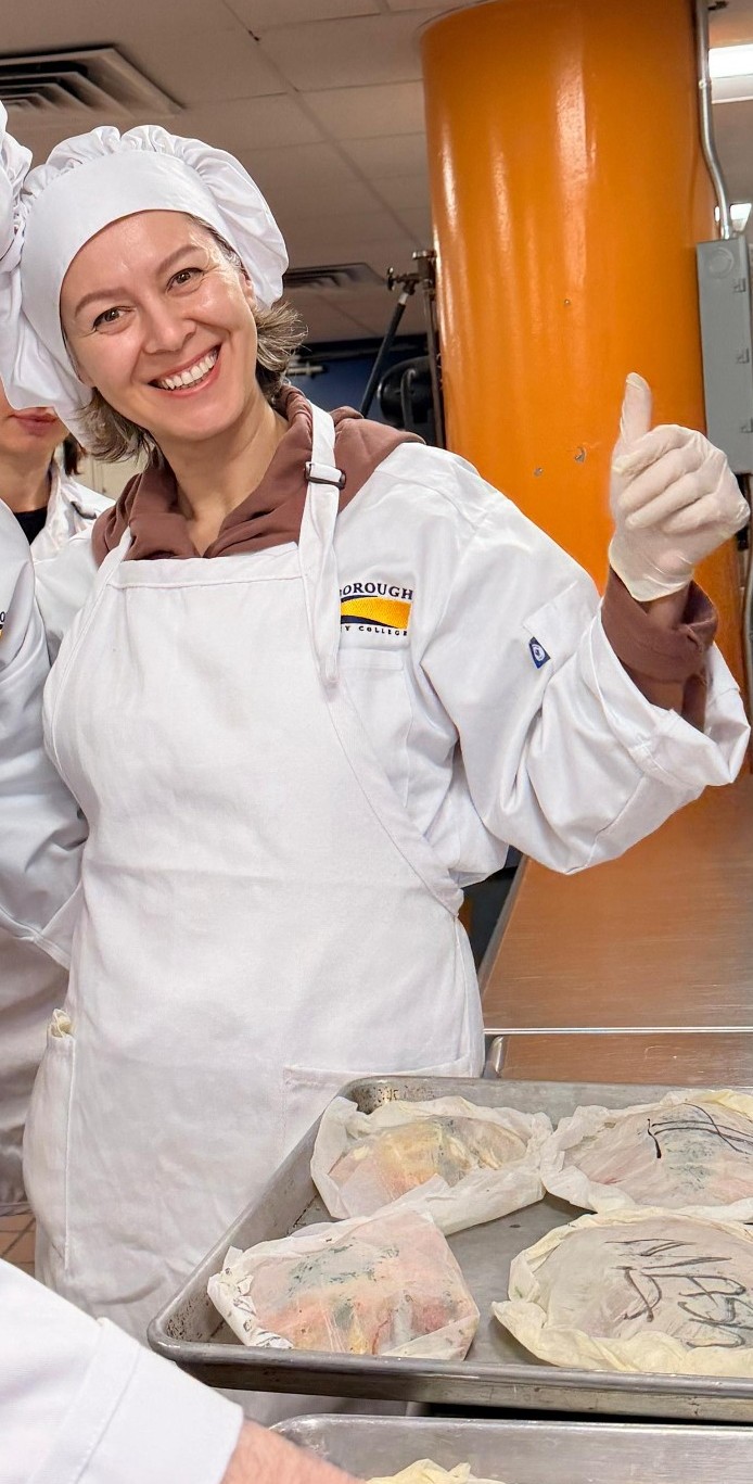 Smiling student in chef's uniform giving a thumbs-up over a tray of wrapped food items in the Kingsborough culinary kitchen.