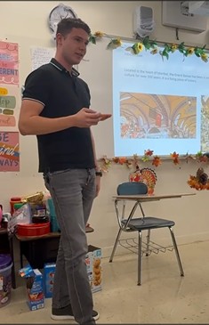 Student in a black polo shirt presenting in front of a classroom, holding a phone, with thte presentation displayed behind on the whiteboard.