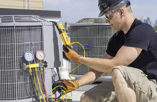 Man in a hard hat and goggles crouching by HVAC equipment on a rooftop.