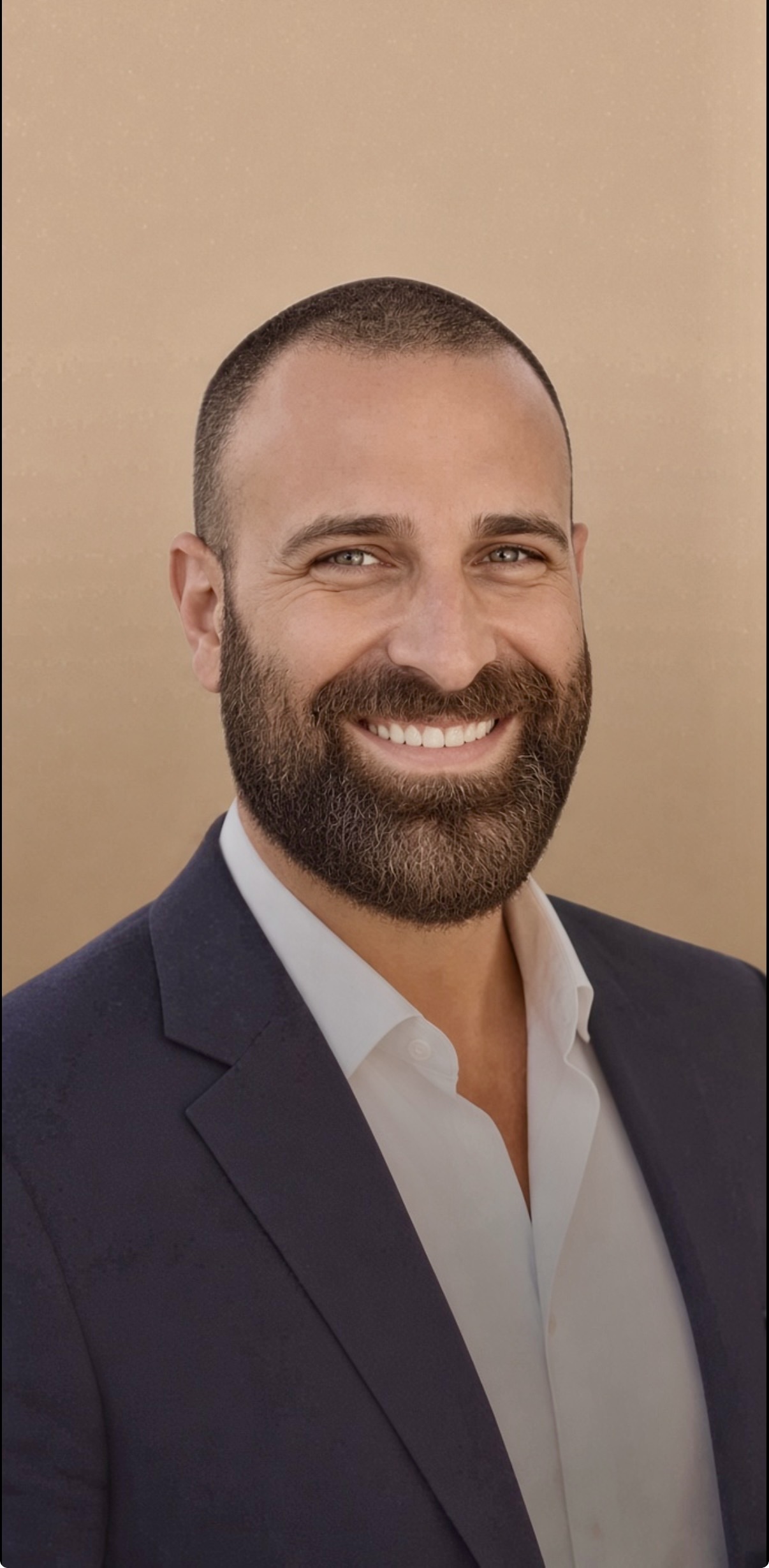 Domenick Brucculeri Smiling young man with a short, light brown beard in a dark blue blazer and white button down shirt