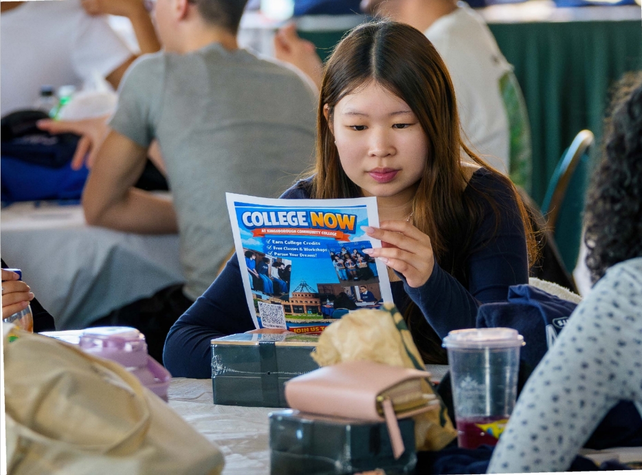 Long haired student sitting at a table looking at a College Now flyer