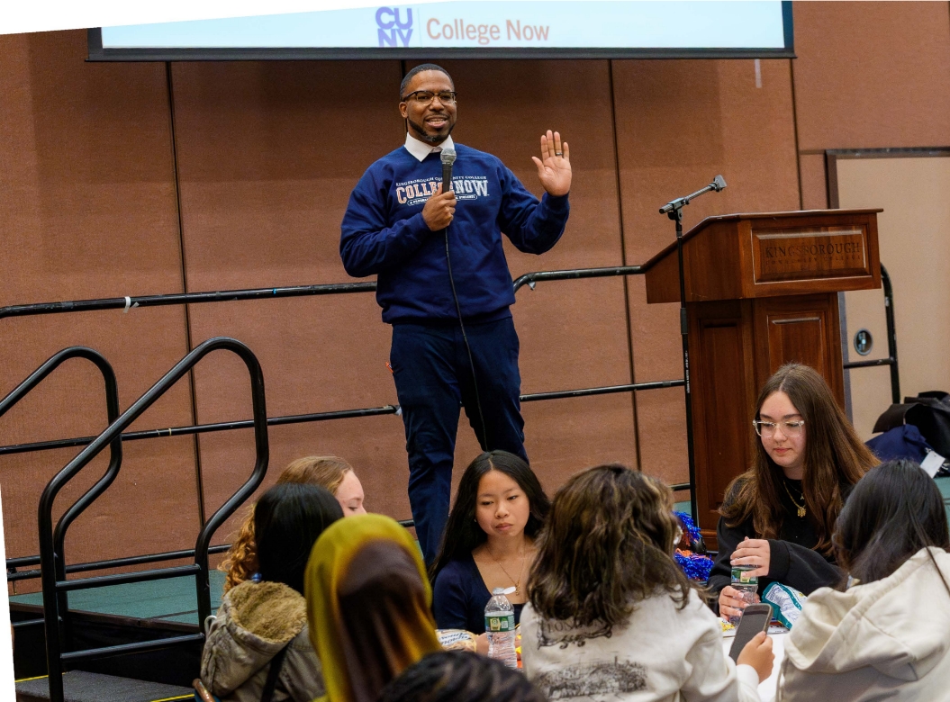 Man in a College Now sweatshirt, holding a mic on stage at an event