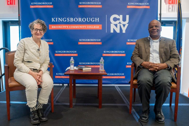 Two people sit in chairs against KCC logo background with a small table between them that has two bottles of water and a book on it. The woman on the left has short, curtly hair and dark-rimmed glasses and is wearing cream-colored top and pants, the man on the right has a shaved head and is wearing a brown blazer with a white button-down and dark slacks.